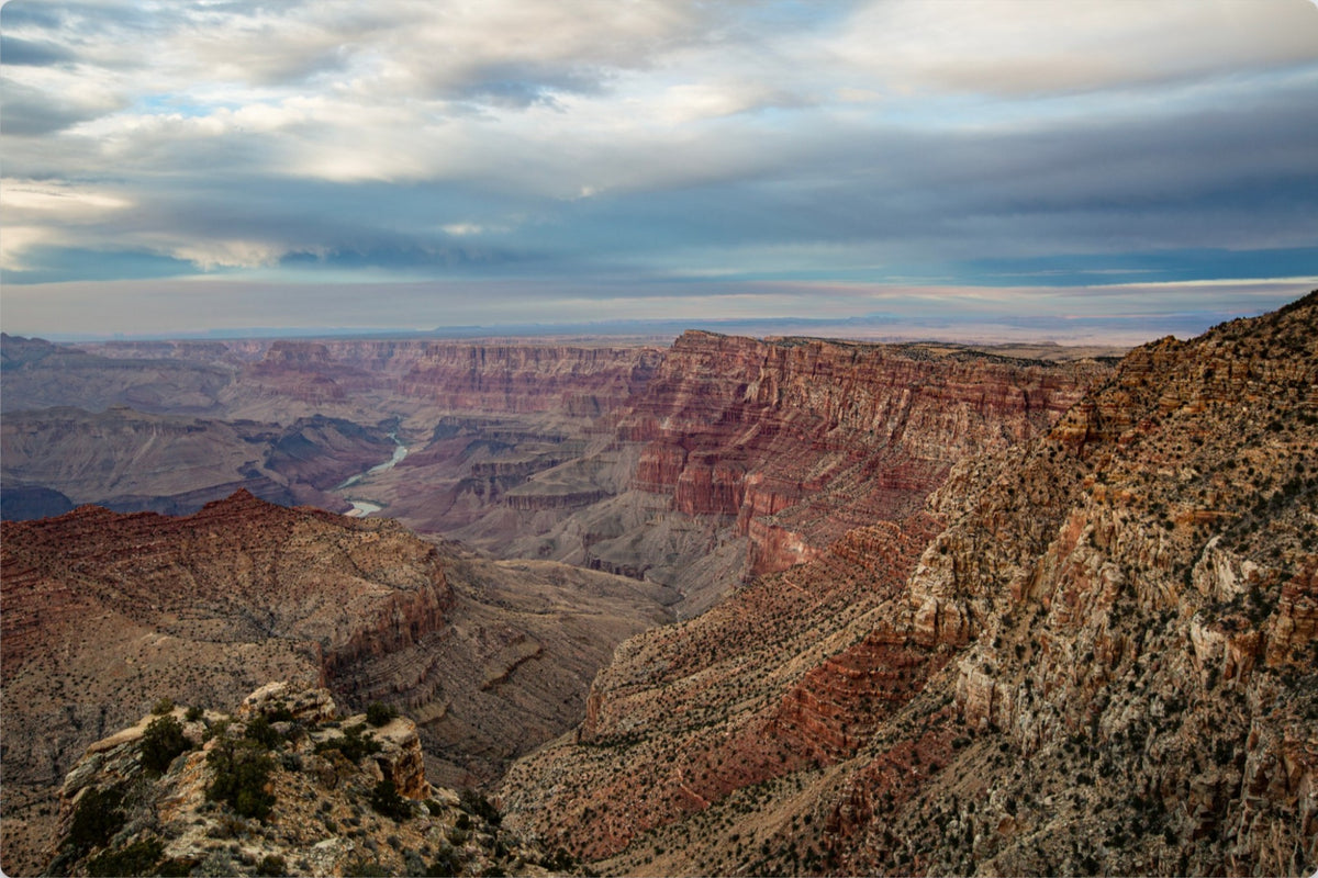 A scenic landscape photograph of the Grand Canyon with layered red and brown rock formations, a winding river in the distance, and a dramatic cloudy sky overhead.